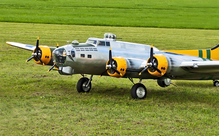 Grauer Modellflieger mit gelben Details und Propellern, der auf einem grünen Feld steht.