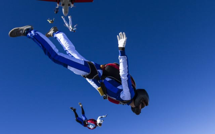 Mehrere Skydiver kurz nach Sprung aus Flugzeug. Die Skydiver tragen weiß-blaue Anzüge und Helme. Der Himmel ist strahlend blau und wolkenlos.