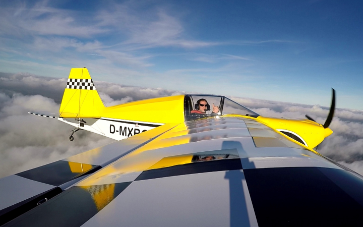 Ultraleichtpilot in Flugzeug in der Luft. Das Flugzeug ist größtenteils gelb. Der Pilot zeigt einen Daumen hoch in die Kamera. Der Himmel ist teilweise bewölkt.