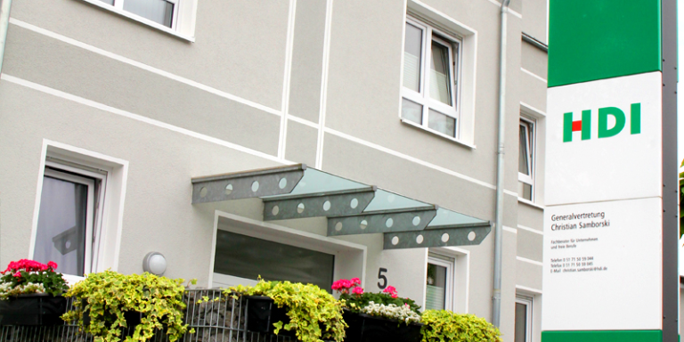 Die agentur befindet sich in einem grauen Gebäude mit der Hausnummer 5. Vor der Agentur befinden dich grüne Pflanzen und Blumen. Außerdem steht ein HDI Schild vor dem Eingang.