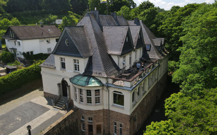 das Verbindungshaus ist weiß und hat ein graues Dach, links neben dem Haus steht ein weiteres Haus und überall sind grüne Bäume