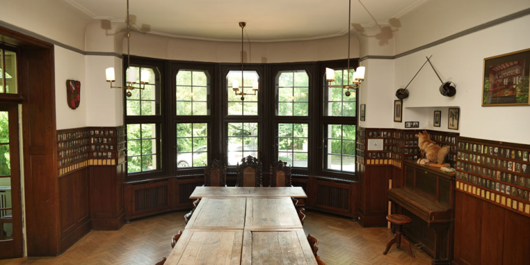 Innenraum des Verbindungshaus mit einer Tafel in der Mitte und Stühle am Ende der Tafel. Hinter den Stühlen ist eine Fensterfront, an den Wänden hängen kleine Bilder und rechts von der Tafel steht ein Klavier auf dem ein ausgestopfter Fuchs steht.