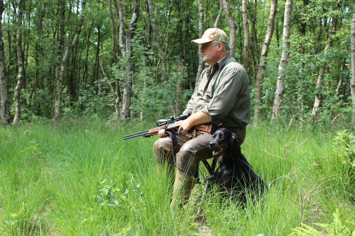 Mann sietzt mit einem Gewehr auf einem Klappstuhl im grünen Wald. Rechts neben Ihm sitzt ein schwarzer Hund