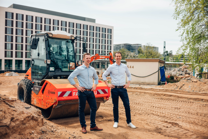 Clemens Reusch und Bernhard Horn auf einer Baustelle, hinter ihnen steht eine Baumschine