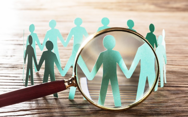 Der Hintergrund ist ein gerillter Holztisch in Nahaufnahme, in der Mitte des Bildes sind aus Papier geschnitzte Menschen, die einen Kreis bilden und sich die Hände reichen. Im Vordergrund des Bildes zeigt eine große Lupe auf einen dieser gebastelten Menschen.