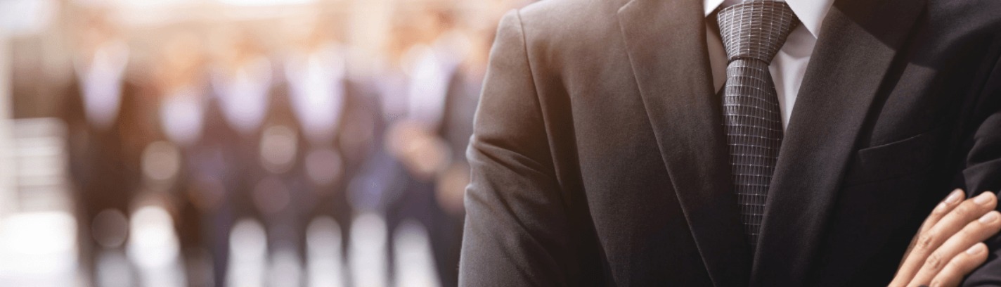 Im Vordergund ist rechts ein schwarzer Anzug mit weißem Hemd zusehen. Im Hintergrund erkennt man verschwommen Männer in schwarzen Anzügen.