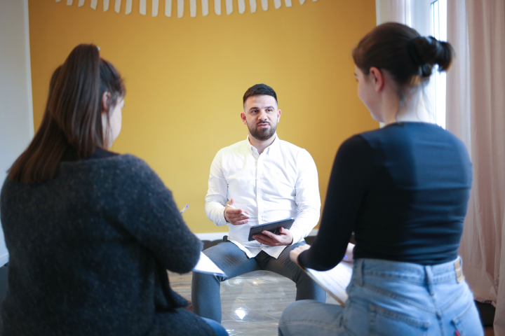 daniel Samloma sitzt  auf einem durchsichtigen Ball und vor einer gelben wand, oben an der wand hängt eine weiße Galande, er hat ein Ipad in der hand und vor Ihm sitzen zwei frauen, die etwas aufschreiben