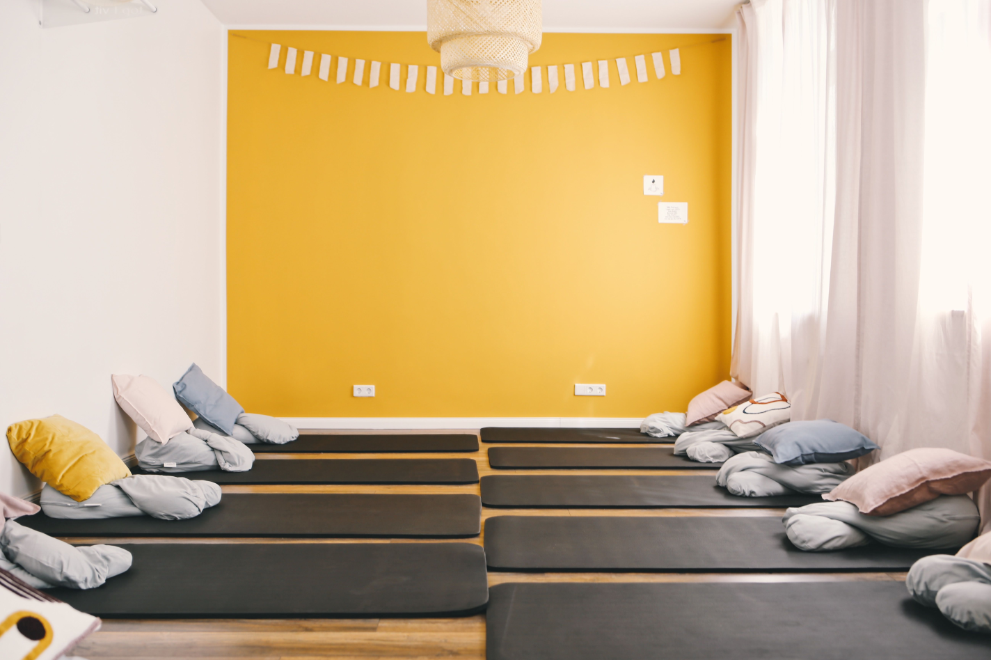 ein großer Raum, geradezu ist eine gelbe Wand mit einer weißen Galande und weißen Steckdosen. An der decke hängt eine beige Lampe. auf dem Boden liegen graue Matten und auf den Matte liegen Kissen, in den Farben rosa, gelb, lila, und weiß