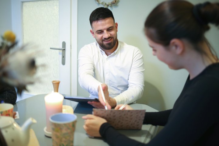 Daniel Salomo und eine Mitarbeiterin sitzen einen Tisch auf dem eine Kerze, eine tasse, eine kanne und einen Blumenstrauß. Die Mitarbeiterin, die rechts außen sitzt, schreibt etwas in ein Heft und Salomo, der links von ihr sitzt, hält ein Ipad in der Hand und zeigt mit seinem Stift auf das heft