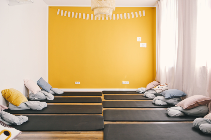 ein großer Raum, geradezu ist eine gelbe Wand mit einer weißen Galande und weißen Steckdosen. An der decke hängt eine beige Lampe. auf dem Boden liegen graue Matten und auf den Matte liegen issen, in den Farben rosa, gelb, lila, und weiß