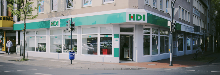 HDI Agentur von außen, die agentur ist weiß, grün und man erkennt das HDI Logo, diese befindet sich im unteren Bereich eines beigen hauses. vor der agentur befindet sich ein gehweg auf dem zwei Ampeln und ein baum stehen. 