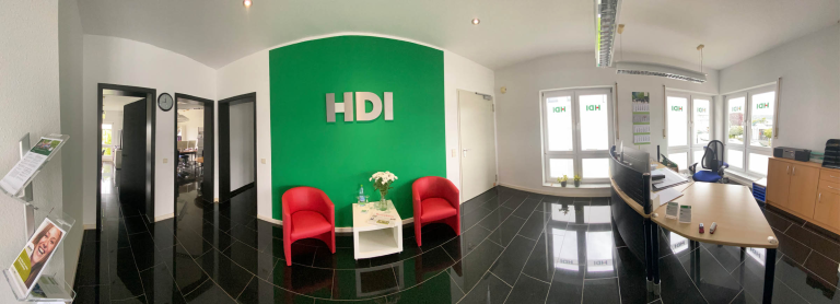 In der Mitte steht ein weißer Tsich mit Blumen. Daneben stehen zwei rote Sessel. Hinter den Möbeln ist eine grüne Wand mit dem silbernen HDI Logo.