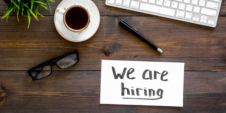Schreibtisch mit Brille, Stift, Kaffeetasse, Pflanze, Tastatur und einem Schild auf dem steht "we are hiring"