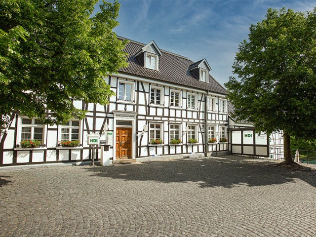 weißes Haus mit grünen Balken und grauem Dach, davor ist ein grauer Platz mit grünen Bäumen