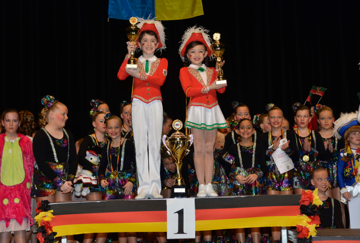 Ein Mädchen und ein Junge stehen auf dem ersten Platz mit jeweils einem Pokal. Dahinter stehen weiter Menschen mit Medallien. 