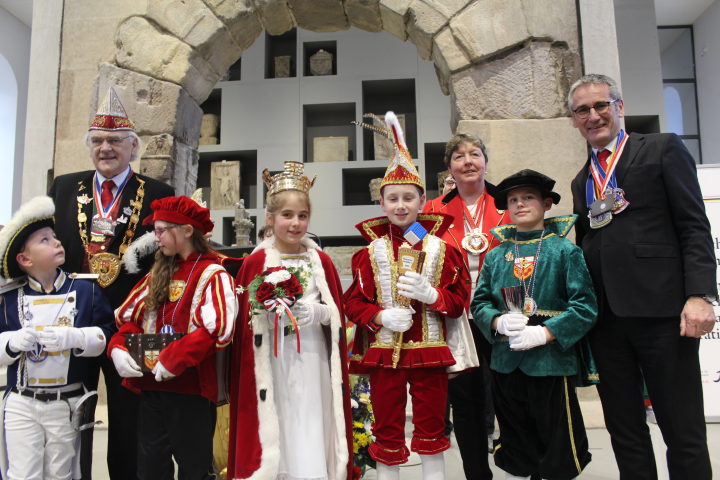 fünf Kinder und drei Erwachsene, die verkleidet vor einem Steinbogen stehen mit Urkunden, Medallien und Blumen. 