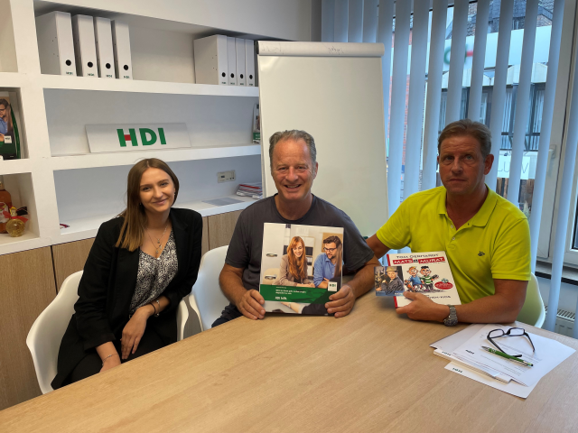 Drei Personen sitzen an einem Tisch. Links ist eine Mitarbeiterin, in der Mitte sitzt Tom Gerhardt und Rechts sitzt Frank Meyer. Gerhardt und Meyer halten HDI Flyer in der Hand. Hinter Ihnen steht ein weißes Regal mit Ordnern und einem HDI Logo. Außerdem steht rechts neben dem Regal eine weiße Tafel.