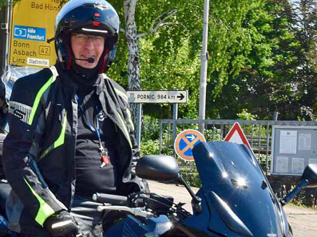 Frank Muthmann auf einem Motorrad, links hinter  ihm ist ein gelbes Schild mit den Aufschrift Bonn, Asbach und Linz, recht von Ihm sind grüne Baume, ein Schild mit der Aufschrift Pornic 984 km und einem Pfeil. dann kommt ein grauer zaun, an diesem ist ein Parken verboten Schild und ein rot, weißes Dreieck. rechts außen ist ein Kasten in diesem Kasten befinden sich Informationen