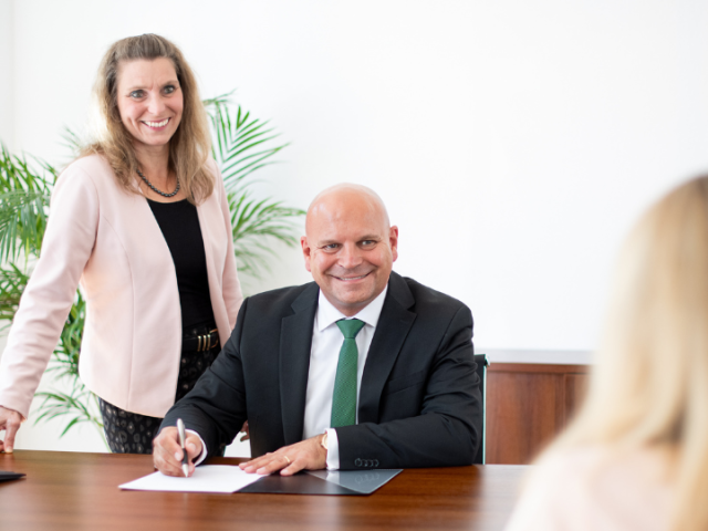 Frank Thatenhorst und Mitarbeiterin in Beratungsgespräch mit Kundin. Er sitzt am Tisch und macht sich Notizen, die Mitarbeiterin steht neben ihm.