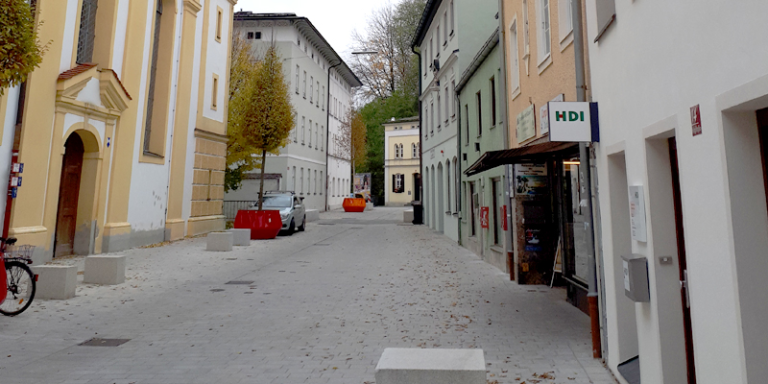 in der Mitte ist ein grauer Gehweg, auf der linken Seite erkennt man den Eingang einer gelben Kirche, dahinter sind gelb, grüne Bäume, dahinter ist ein hellgrünes Haus und ein weißes Haus . Am Ende des Gehweges steht ein gelbes Haus. Auf der rechten Seite des Gehweges sieht man die HDI Agentur, die in einem Haus ist, danach kommt ein oranges Haus, ein grünes Haus und ein weißes Haus