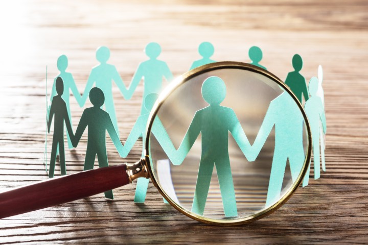 Der Hintergrund ist ein gerillter Holztisch in Nahaufnahme, in der Mitte des Bildes sind aus Papier geschnitzte Menschen, die einen Kreis bilden und sich die Hände reichen. Im Vordergrund des Bildes zeigt eine große Lupe auf einen dieser gebastelten Menschen.