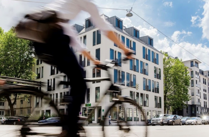 Fahrradfahrer verschwommen und im Hintergrund ein weißes Haus mit Fenstern und unten ist die HDI Agentur