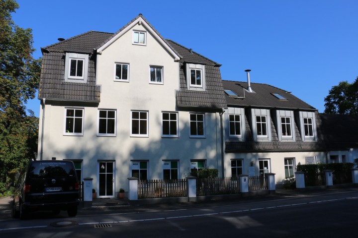 beiges Haus mit blauen Himmel und links steht ein schwarzes Auto , vor dem Haus ist ein brauner Zaun