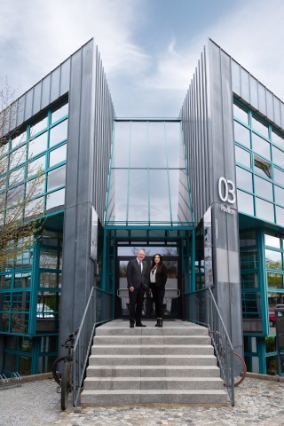 Hans Wolff und Mitarbeiterin stehen auf einer Treppe vor einem Gebäude mit großer Glasfront.