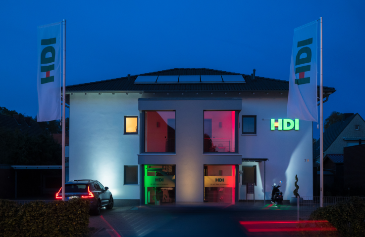 Agentur bei Nacht, die Fenster sind rot beleuchtet, das HDI Logo rechts an der hauswand leuchtet grün. links vor der Haus steht ein Auto und vor dem weißen Haus stehen links und rechts Werbefahnen