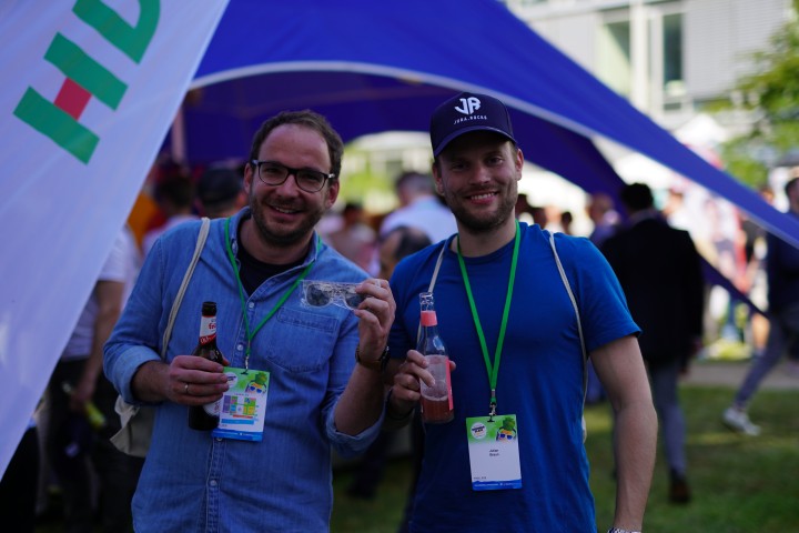 Zwei Männer in blauen Shirts schaune lächelnd in die Kamera. Beide haben ein Getränk in der Hand und tragen einen Veranstaltungsausweis um den Hals.