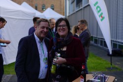 Bild von einer Frau und einem Mann die lächelnd in die Kamera schauen. Im Hintergrund sieht man einen Aufsteller mit dem HDI Logo.