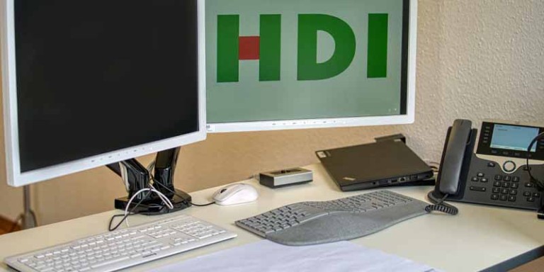Schreibtisch mit zwei Bildschirneb, Tastaturen und einem Telefon. Auf dem rechten Bildschirm wird das HDI Logo angezeigt.
