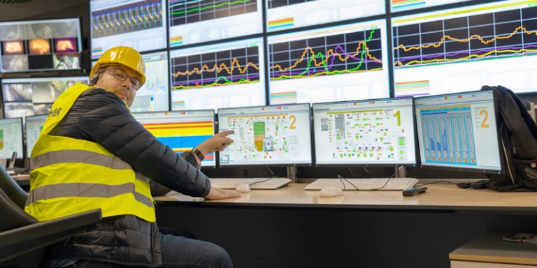Bild von Mitarbeiter in gelber Warnweste und mit Schutzhelm. Er deutet auf Bildschirme mit Grafiken.