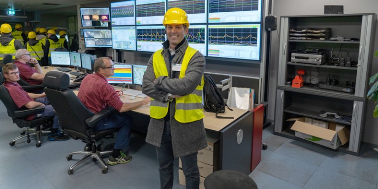 Bild von Mitarbeitern an Schreibtischen. Vorne steht ein Mann in Warnweste mit Schutzhelm und schaut lächelnd in die Kamera.