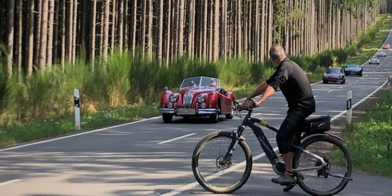 rechts steht ein Fahrradfahrer, der vor einer Straße steht, diese Straße wird von sechs Autos befahren, diese Autos sind rot, lila, blau, schwarz, weiß und orange. neben der Straße sind auf beiden Seiten grüne Bäume und grüne Büsche