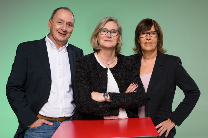 gruppenfoto mit drei Mitarbeitern, diese stehen hinter einem roten Tisch und vor einer grünen Wand