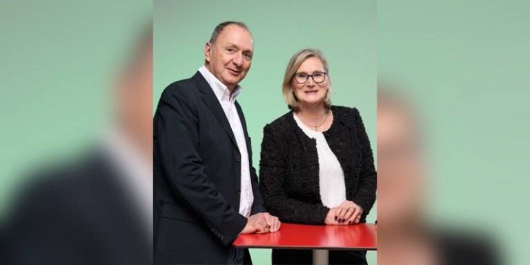 Gruppenfoto mit Heike Schlick-Karst rechts und einem Mitarbeiter links, diese stehen hinter einem roten Tisch und vor einer grünen Wand