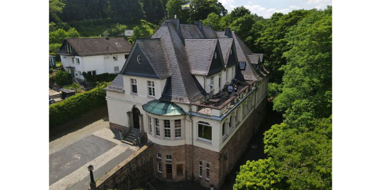 das Verbindungshaus ist weiß und hat ein graues Dach, links neben dem Haus steht ein weiteres Haus und überall sind grüne Bäume