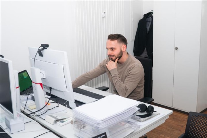 Ein Mitarbeiter sitzt an seinem Schreibtisch und arbeitet an seinem PC. Rechts neben dem Pc steht ein Laptop und links von seinem PC ist eine Aufbewahrungsbox für Dokumenten.