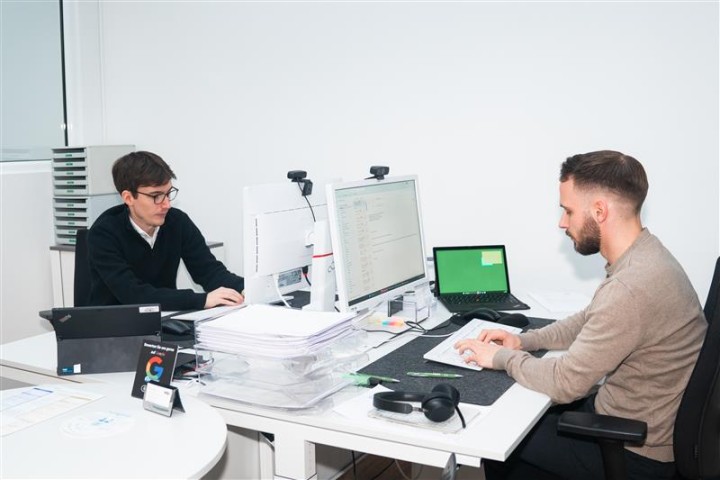 Ein Mitarbeiter sietzt an einem Schreibtisch, auf dem ein Pc und ein Laptop stehen. Dabei arbeitet er an seinem PC.