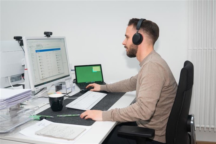 Ein Mitarbeiter sitzt an seinem Schreibtisch und arbeitet an seinem PC. Rechts neben dem Pc steht ein Laptop und er trägt auf deinem Kopf ein Headset.