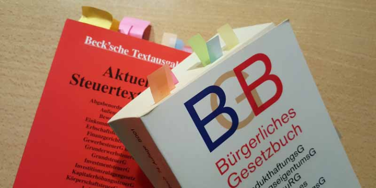ein Buch ist rot und über steuern, diese liegt auf einem brauen Tisc und darüber leigt ein weißes Buch über Recht. beide haben bunte Post its, als Makierung im Buch