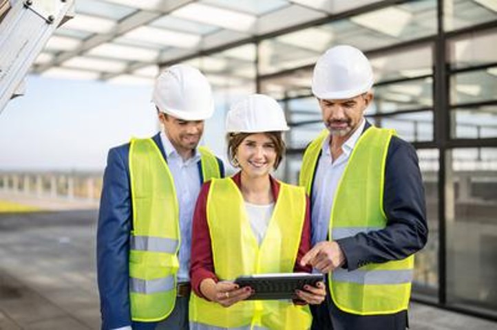 Drei Menschen haben gelbe Westen und weiße Helme an. Eine Frau, die lächelnd in die Kamera guckt, steht in der Mitte und hat ein Ipad in der Hand. Links und rechts steht jeweils ein Mann neben der Frau. Die beiden Männer gucken auf das Ipad.