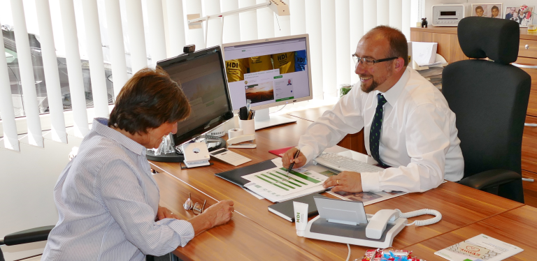 Karl Kramer sitzt hinter dem Schreibtisch auf einem Stuhl vor einem PC, vor Ihm sitzt eine Mitarbeiterin und Sie führen ein Gespräch