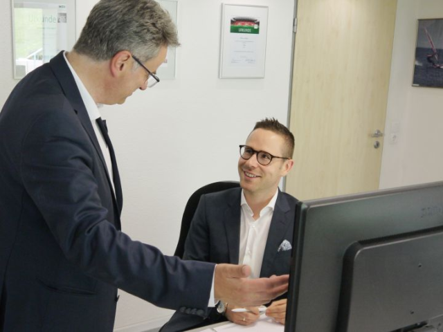 Auf der linken Seite steht der Agenturleiter Marcus Ailinger und unterhält sich gerade mit seinem Kollegen. Beide Herren tragen eine Brille und einen Anzug.