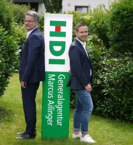 Auf der linken Seite steht der HDI Agenturleiter Marcus Ailinger seitlich und lächelt in die Kamera. Neben ihm ist ein umgedrehtes Eingangsschild seiner Agentur mit dem HDI-Logo in grün, weiß, rot. Rechts von dem Schild steht seitlich angelehnt sein Kollege, der ebenfalls in die Kamera lächelt. Beide tragen einen dunkelblauen Anzug. Das Bild wurde in einem Garten mit vielen grünen Bäumen geschossen.
