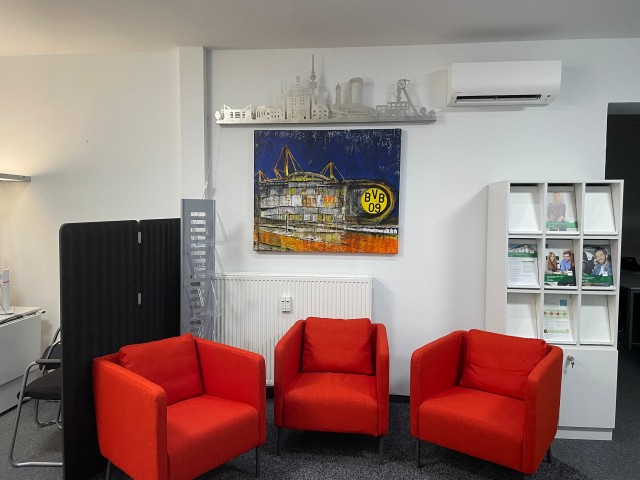 In dem Büro erkennt man in der Mitte eine Sitzecke mit drei roten Sessel und dahinter hängt ein selbstgemaltes Bild mit der BVB Zentrale. 