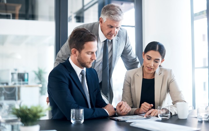 Zwei Männer mittleren Alters und eine Frau unterhalten sich geschäftlich. Alle drei haben einen Anzug an, die beiden Männer mit Krawatte. Der linke Mann hat braunde Haare und einen Bart, der Mann in der Mitte hat graues Haar und kein Bart. Die Frau links ist geschminkt und trägt einen Zopf mit langen schwarzen Haaren.
