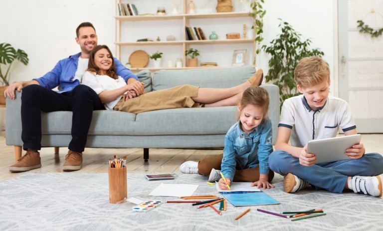 Vater und Mutter liegen zusammen auf der Couch und beobachten ihre beiden Kinder fröhlich beim Spielen. Die Tochter malt mit Buntstiften, der Junge spielt am Tablet.