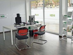 In dem Büro steht ein Schreibtisch auf dem ein PC steht. Hinter dem Schreibtisch steht ein schwarzer Stuhl und vor dem Tisch stehen zwei rote Stühle. Rechts in der Ecke steht ein Aufsteller mit HDI Zeitschriften. Außerdem sieht man eine Fensterfront